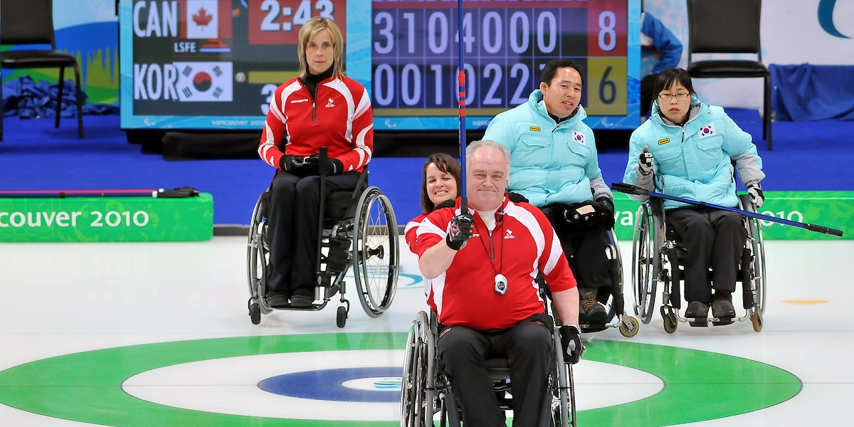 Jim Armstrong curling with Ina Forrest holding his chair and Sonia Gaudet in the background on the ice