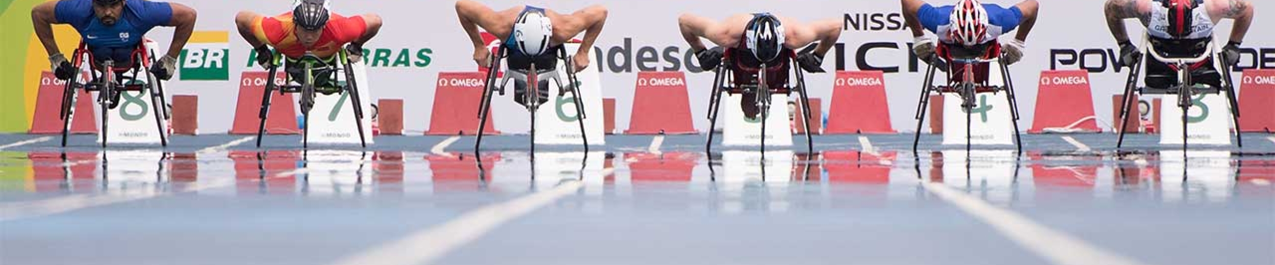 Racers line up in their wheelchairs at the 100m start line; 6 racers side by side in their lanes