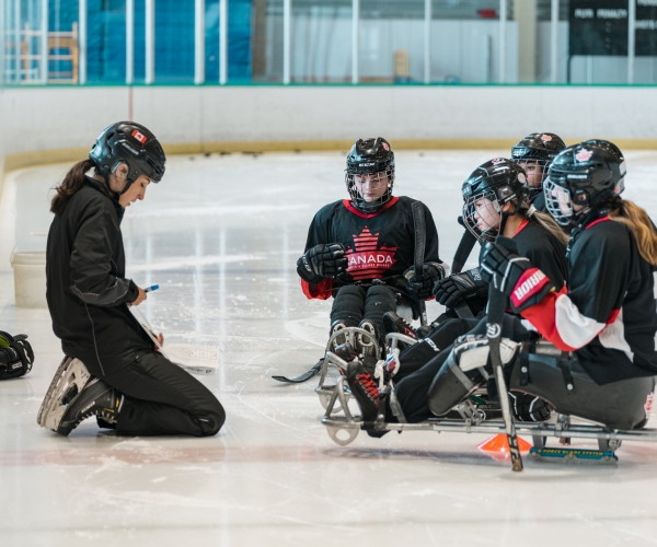 A group of female Para ice hockey players huddle around their coach on the ice. 