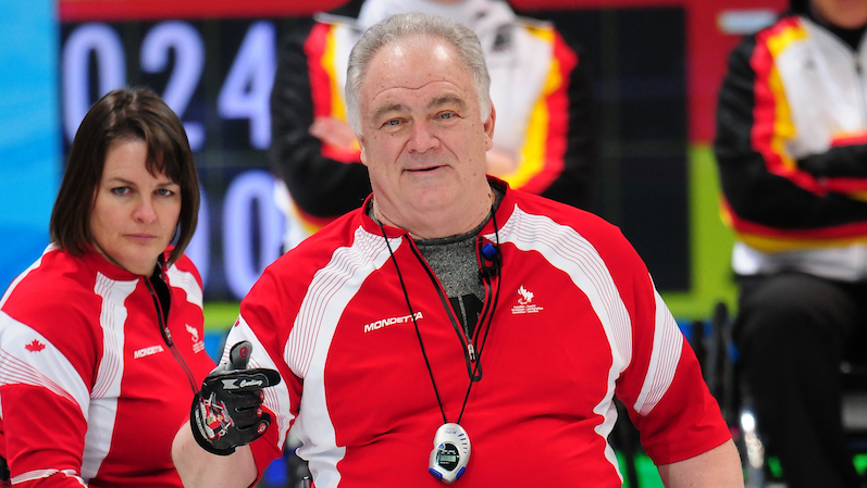 Jim Armstrong looks on with a satisfied semi-smile. Wearing a red shirt with white stripes.