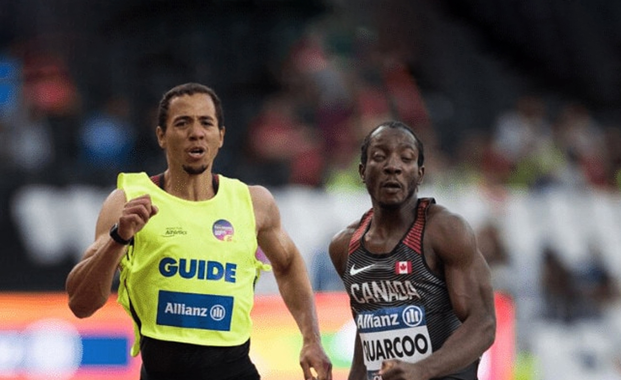 Canadian Para athlete George Quarcoo sprints alongside his sighted guide during a race. Quarcoo wears a black Team Canada uniform with red accents, displaying his name and a Canadian flag on his chest. His guide, dressed in a bright yellow vest labeled "GUIDE," runs beside him. Both athletes show intense focus as they compete in a stadium with a blurred crowd in the background.