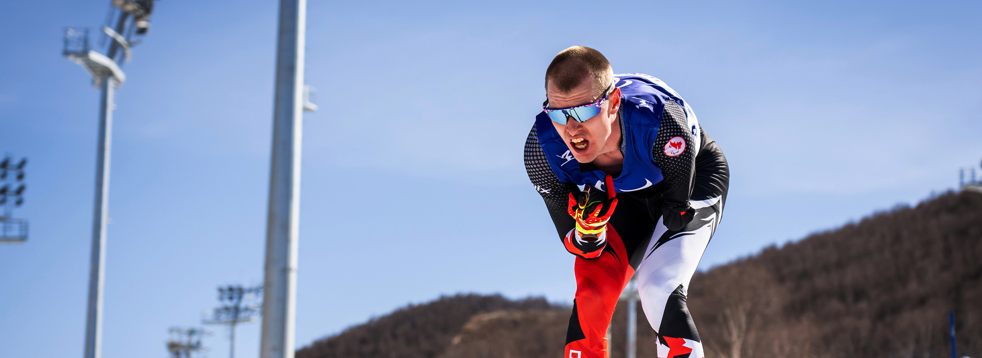 Canadian Para nordic skier Mark Arendz competes in a biathlon event at the Paralympic Winter Games, pushing hard on his skis while wearing a blue and black race suit with red gloves.