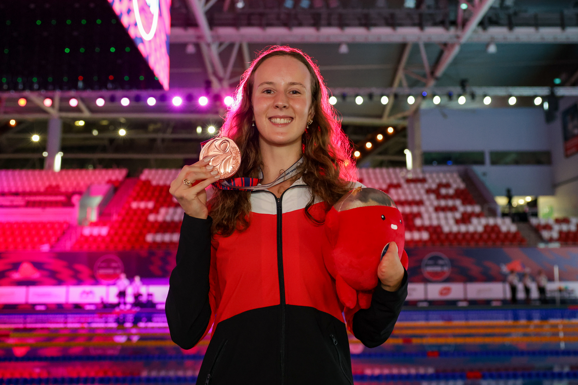 A smiling athlete stands inside a brightly lit indoor pool venue, holding a bronze medal in one hand and a red plush mascot in the other. She wears a red, white, and black team jacket, and colourful lights illuminate the background stands and pool.