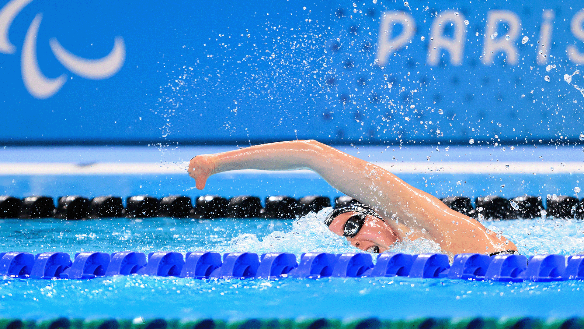 A Para swimmer in goggles and a swim cap powers through a freestyle race, her arm extended mid stroke as water splashes around her. Blue lane markers and a Paris 2024 backdrop frame the scene.