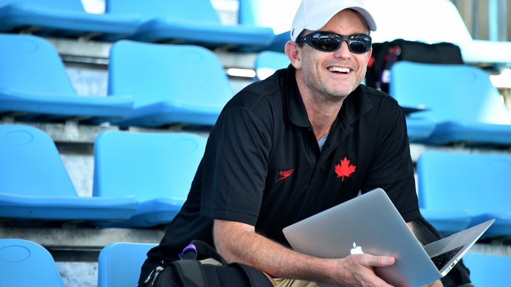 Coach Spotlight on Wayne Lomas, Wayne sits in a set of bleachers with a big smile on his face