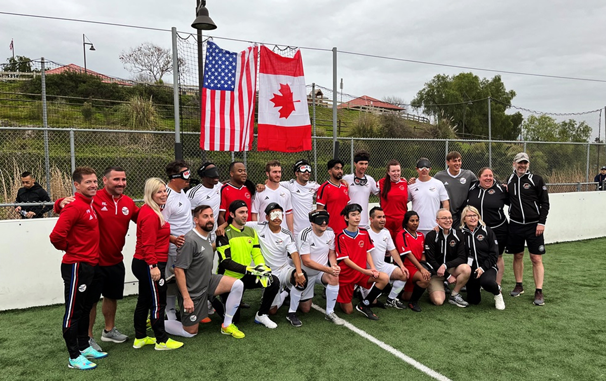 Canadian and U.S. blind football (Football 5-a-side) players and staff pose together for a group photo on the field after a match. Both the Canadian and American flags hang behind them.