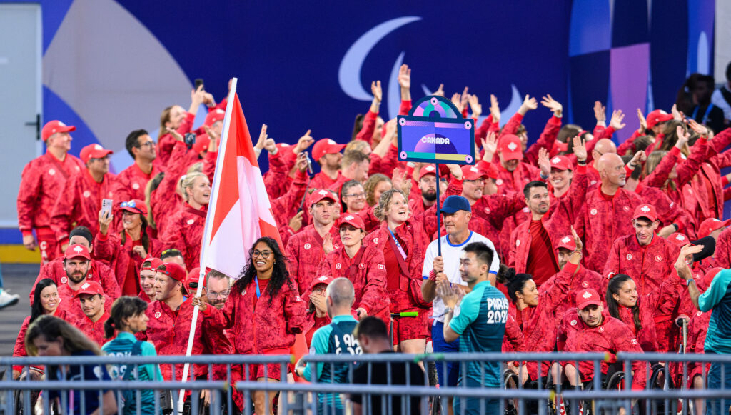 The Canadian Paralympic Team, dressed in red uniforms, enters the stadium at the Opening Ceremony of the Paris 2024 Paralympic Games. Team members wave and cheer, with the Canadian flag and team sign held proudly at the front.