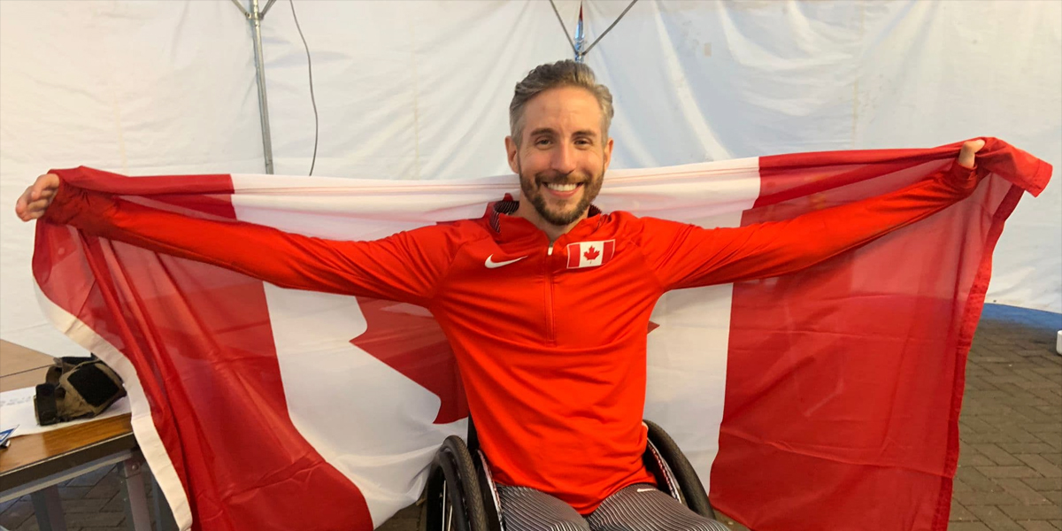 Canadian Wheelchair racer Cody Fournie celebrates following a race by holding up the Canadian flag behind him with his arms outstretched holding the flag.