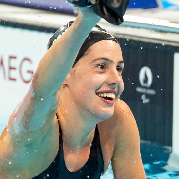 Canadian Para swimmer Aurélie Rivard smiles with joy as she raises her arm in celebration after finishing a race at the Paralympic Games. She is still in the pool, water dripping from her, wearing a black swim cap and swimsuit. The background shows the timing system with "Omega" visible.