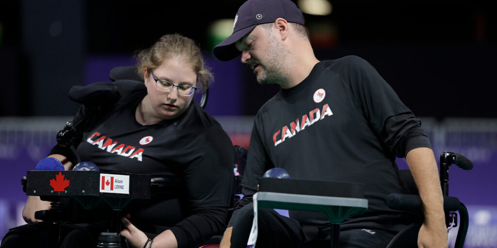 Canadian boccia athlete Alison Levine, wearing a black Canada team shirt, is seated in her wheelchair and is focused on her boccia ramp. A coach or assistant, also in a black Canada shirt, leans over to assist or give advice during the match. Several boccia balls are visible on the ramp, and both individuals are concentrating on the game.