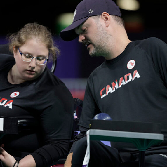 Canadian boccia athlete Alison Levine, wearing a black Canada team shirt, is seated in her wheelchair and is focused on her boccia ramp. A coach or assistant, also in a black Canada shirt, leans over to assist or give advice during the match. Several boccia balls are visible on the ramp, and both individuals are concentrating on the game.