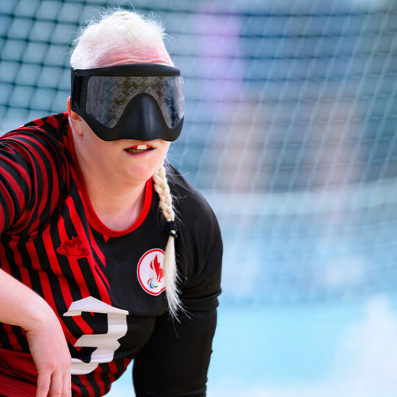 Canadian goalball player Whitney Bogart is seen in action during a match. She wears a black and red team jersey with the number 3, along with a black eye mask, which is standard equipment in goalball. Whitney is crouching on the court, focused and ready to defend.