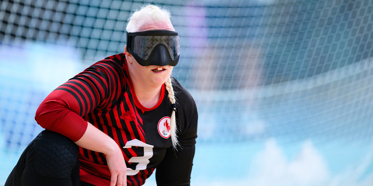 Canadian goalball player Whitney Bogart is seen in action during a match. She wears a black and red team jersey with the number 3, along with a black eye mask, which is standard equipment in goalball. Whitney is crouching on the court, focused and ready to defend.