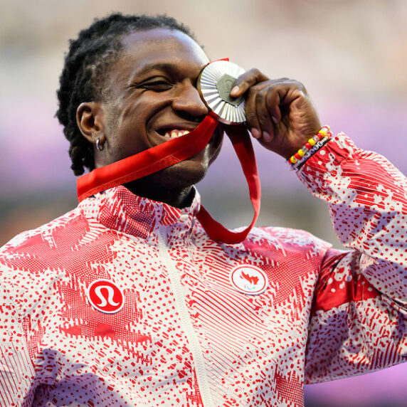 The image shows Jesse Zesseu, a Canadian Para-athlete, smiling with pride as he bites his silver medal during the medal ceremony at the Paris 2024 Paralympic Games. He is wearing a red and white patterned jacket with the Canadian Paralympic Team and Lululemon logos visible. Zesseu is holding the medal with one hand, and the red ribbon hangs down. The background is blurred, emphasizing his joyful expression.