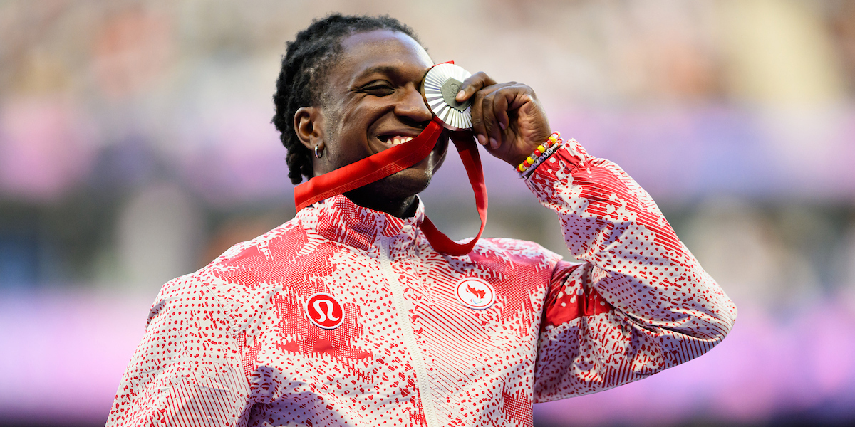 The image shows Jesse Zesseu, a Canadian Para-athlete, smiling with pride as he bites his silver medal during the medal ceremony at the Paris 2024 Paralympic Games. He is wearing a red and white patterned jacket with the Canadian Paralympic Team and Lululemon logos visible. Zesseu is holding the medal with one hand, and the red ribbon hangs down. The background is blurred, emphasizing his joyful expression.