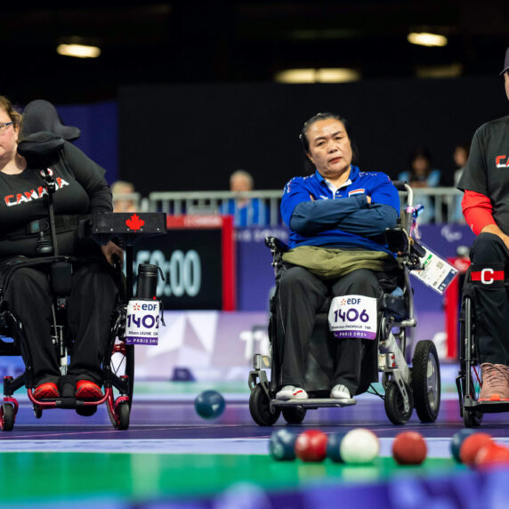 Alison Levine throws a blue ball while Iulian Ciobanu and an opponent watch in front of a set of balls during a match of Boccia.