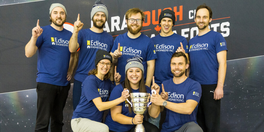 A group of eight people wearing matching blue Edison Engineers t-shirts and winter hats are posing together with a large trophy. They are smiling and holding up one finger to indicate they are number one, celebrating their victory at the ParaTough Cup event.