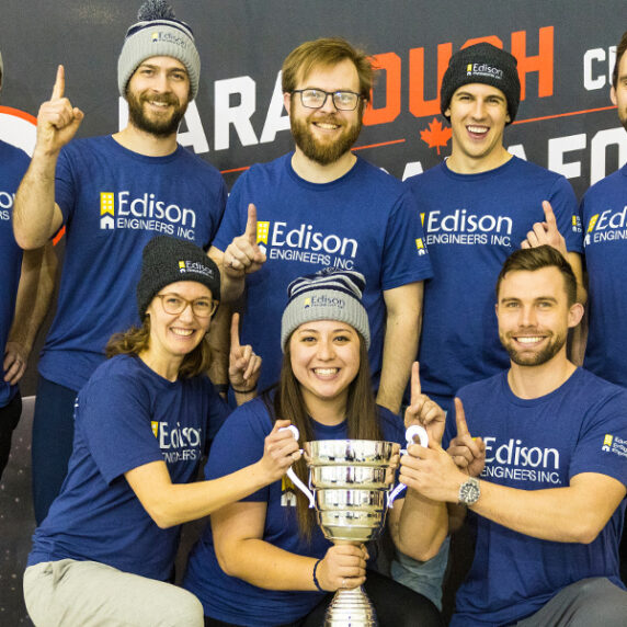 A group of eight people wearing matching blue Edison Engineers t-shirts and winter hats are posing together with a large trophy. They are smiling and holding up one finger to indicate they are number one, celebrating their victory at the ParaTough Cup event.