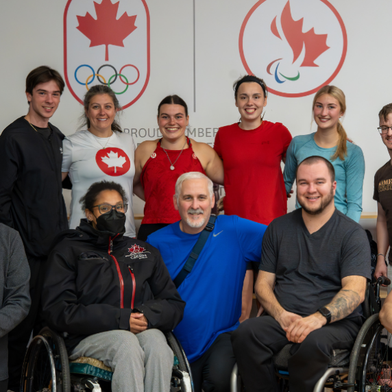 The image shows a group of diverse individuals, some standing and some in wheelchairs, posing together in front of a wall displaying the logos of the Canadian Olympic and Paralympic teams. They are all smiling, with a positive and inclusive atmosphere. The setting appears to be a training or team-building facility, reflecting a strong sense of camaraderie and teamwork.