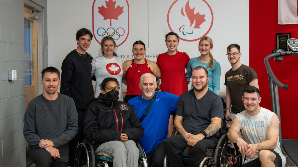 The image shows a group of diverse individuals, some standing and some in wheelchairs, posing together in front of a wall displaying the logos of the Canadian Olympic and Paralympic teams. They are all smiling, with a positive and inclusive atmosphere. The setting appears to be a training or team-building facility, reflecting a strong sense of camaraderie and teamwork.