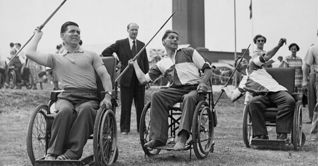 Black and white photo of wheelchair athletes participating in a javelin event, with three men in wheelchairs preparing to throw javelins in an outdoor setting, observed by spectators in the background.