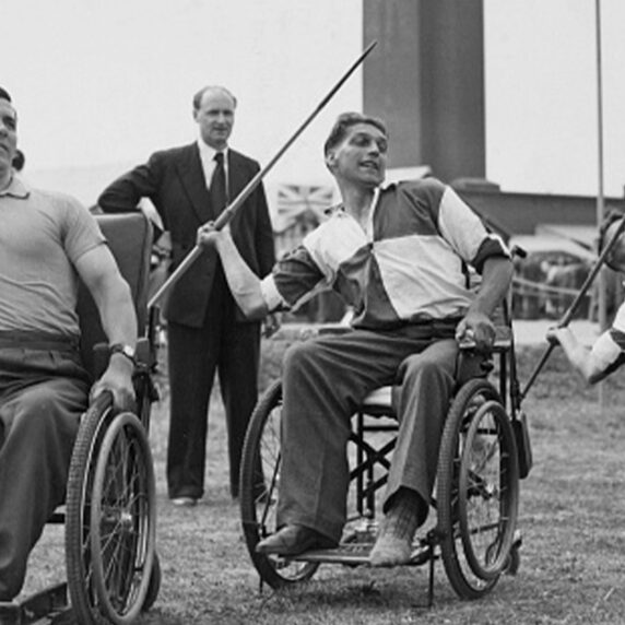 Black and white photo of wheelchair athletes participating in a javelin event, with three men in wheelchairs preparing to throw javelins in an outdoor setting, observed by spectators in the background.