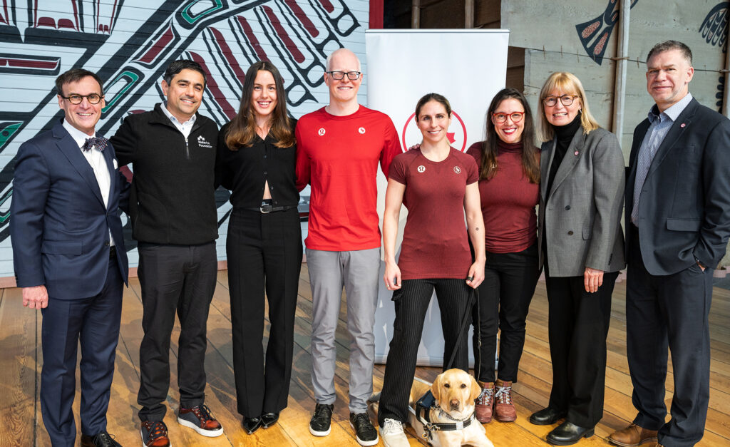 A group of eight people, including Paralympic athletes, foundation representatives, and a service dog, stands side by side, smiling at the camera. The setting appears to be indoors with a backdrop featuring Indigenous artwork. The group includes men and women dressed in both formal and athletic wear, highlighting a partnership or community event. A yellow labrador service dog sits calmly at the feet of one of the individuals.