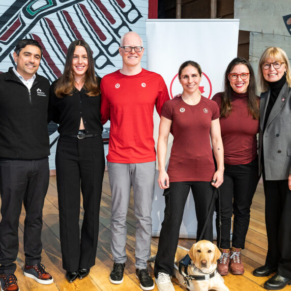 A group of eight people, including Paralympic athletes, foundation representatives, and a service dog, stands side by side, smiling at the camera. The setting appears to be indoors with a backdrop featuring Indigenous artwork. The group includes men and women dressed in both formal and athletic wear, highlighting a partnership or community event. A yellow labrador service dog sits calmly at the feet of one of the individuals.