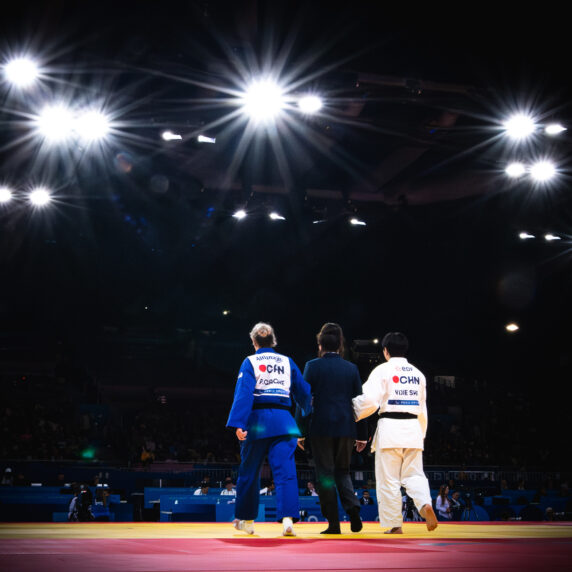 A visually impaired judoka wearing a blue gi, guided by a coach and official, walks towards the center of a brightly lit competition arena under a spotlight, with the audience in the background.