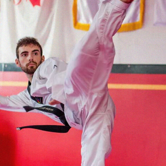 Canadian Para taekwondo athlete Anthony Cappello performs a high kick in a dojo, wearing a white taekwondo uniform and a black belt. The Canadian and Quebec flags are visible in the background, representing his Canadian roots.