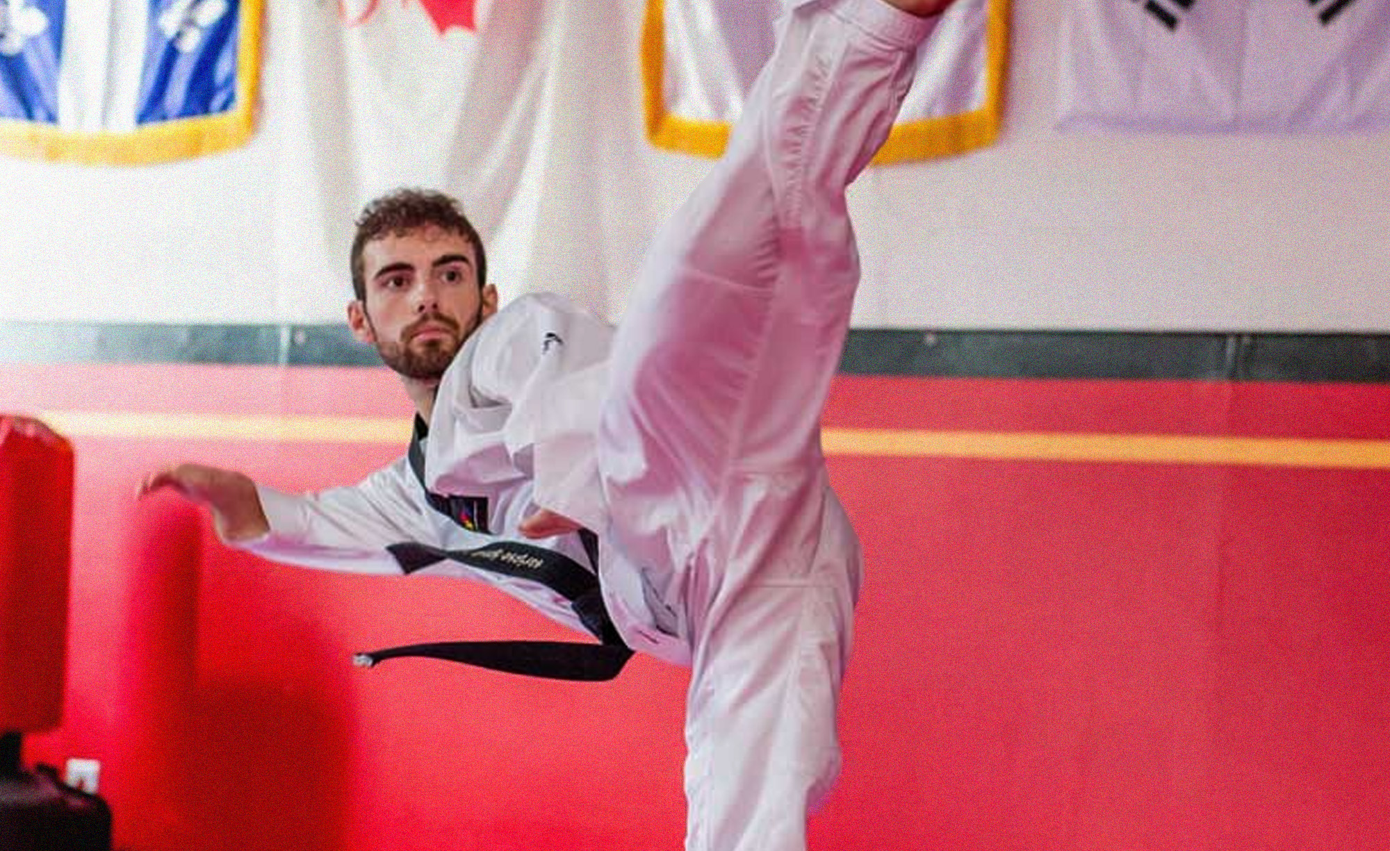 Canadian Para taekwondo athlete Anthony Cappello performs a high kick in a dojo, wearing a white taekwondo uniform and a black belt. The Canadian and Quebec flags are visible in the background, representing his Canadian roots.