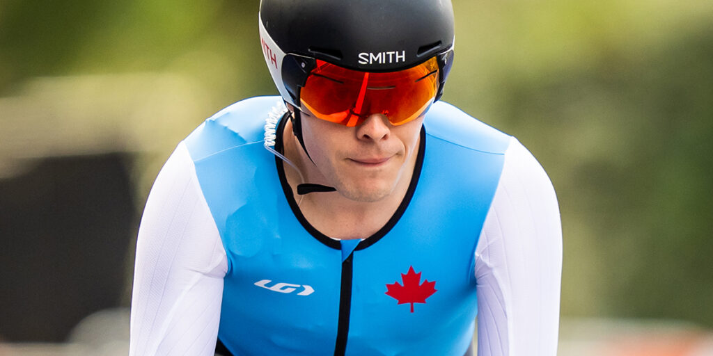 Canadian Paralympian Nathan Clement races on his bike wearing a light blue and white cycling suit featuring a red maple leaf. His focused gaze and aerodynamic helmet emphasize his determination during the competition.