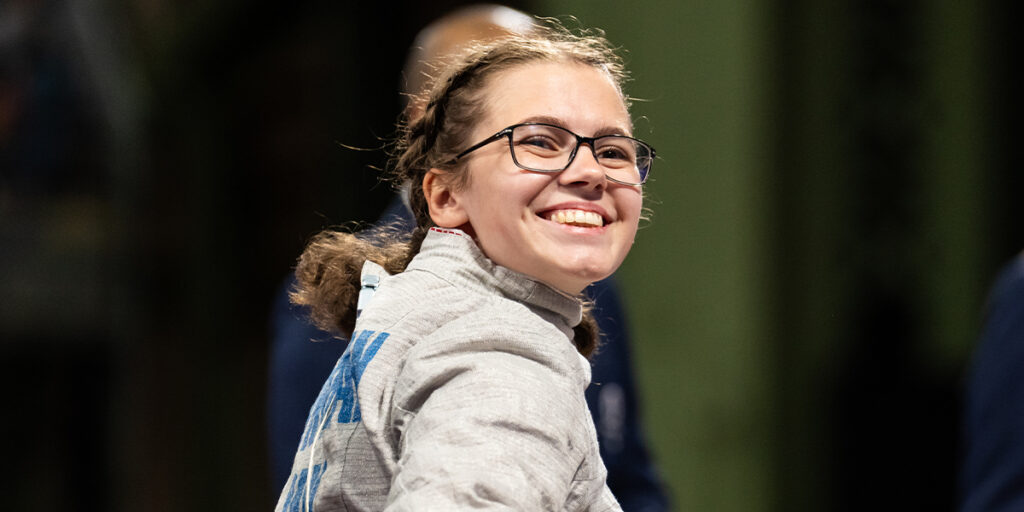 Canadian Paralympian Trinity Lowthian smiles confidently after a wheelchair fencing bout, wearing her silver fencing jacket and glasses, with her braided hair neatly tied back. The bright atmosphere highlights her celebratory moment.