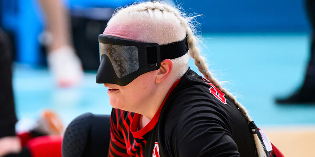 Canadian Paralympian Whitney Bogart competes in a goalball match, wearing a black and red jersey with a blindfold as she focuses on the game. Her braided hair adds a personal touch to her determined expression.