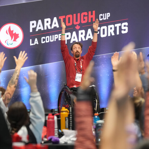 Canadian Paralympian Josh Dueck leads a group of participants in a pre-event stretching routine while emceeing a ParaTough Cup event. Seated in his wheelchair, Josh raises his arms high, energizing the crowd against the backdrop of a banner reading “ParaTough Cup / La Coupe ParaForts.”