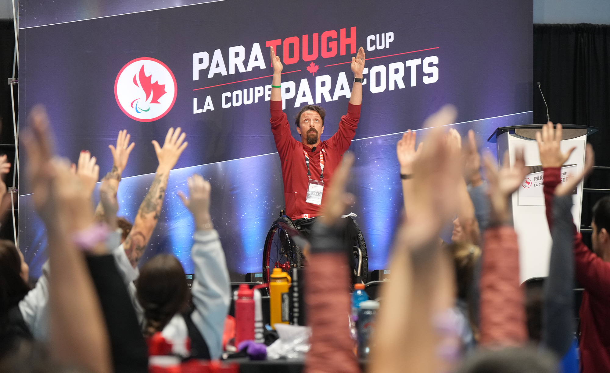 Canadian Paralympian Josh Dueck leads a group of participants in a pre-event stretching routine while emceeing a ParaTough Cup event. Seated in his wheelchair, Josh raises his arms high, energizing the crowd against the backdrop of a banner reading “ParaTough Cup / La Coupe ParaForts.”