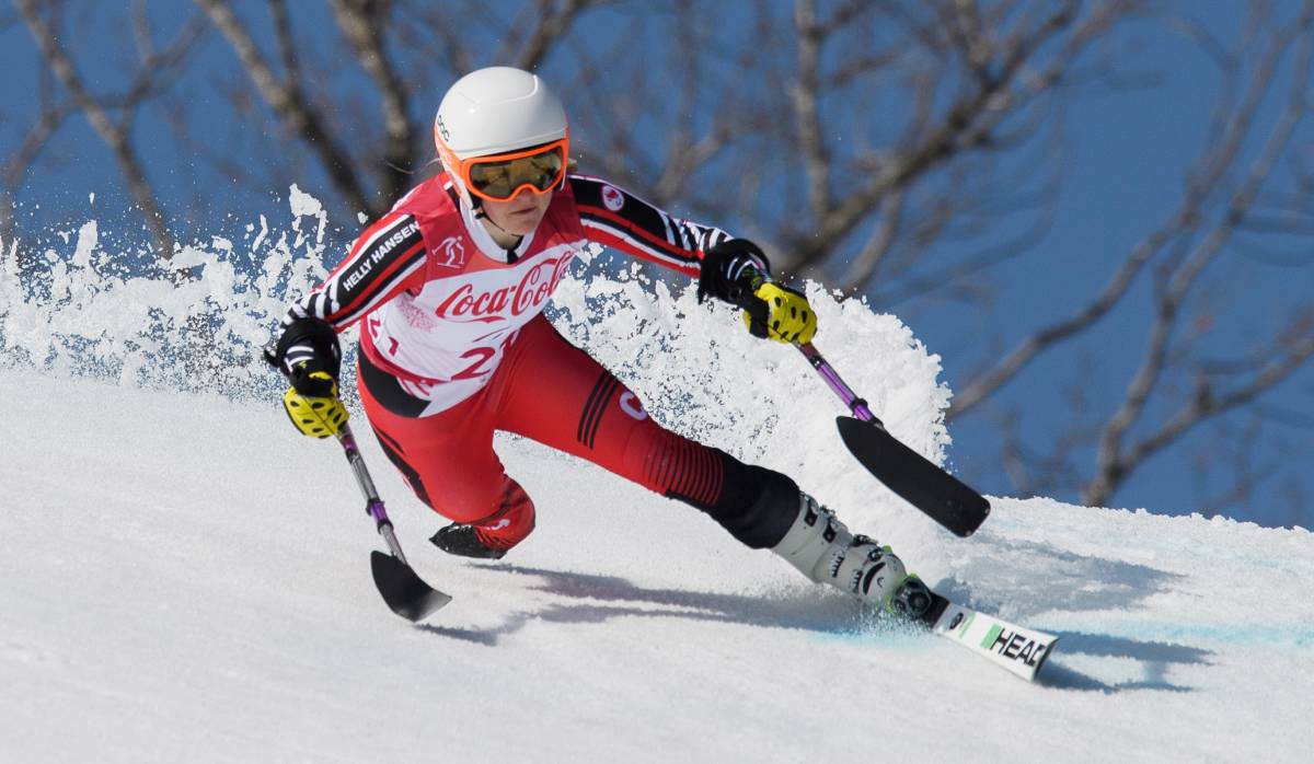 Canadian Paralympian Frédérique Turgeon competes in alpine skiing on a snowy slope, wearing a red and white racing suit, a white helmet, and yellow gloves. She uses outriggers for balance and speed as she carves through the snow.