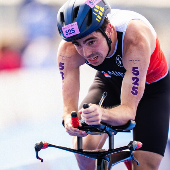 A determined athlete in a Canadian triathlon suit races on a specialized bike during the Paris 2024 Paralympic Games. The number 525 is visible on their arms and helmet, emphasizing their competitive spirit.