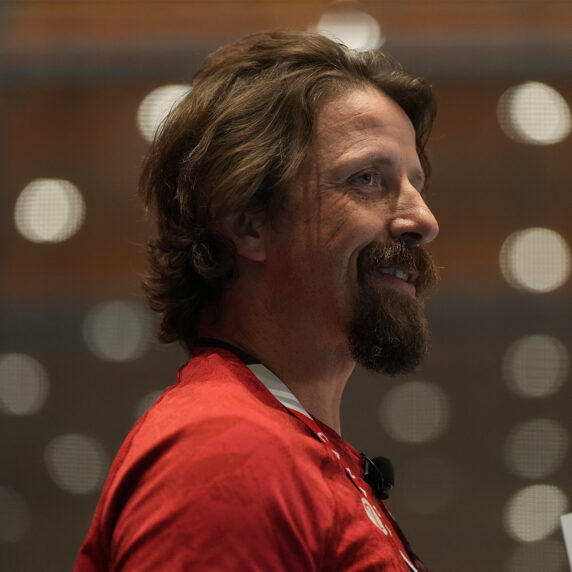 Profile view of Canadian Paralympic gold medallist Josh Dueck smiling, wearing a red shirt and speaking on stage with a microphone headset.