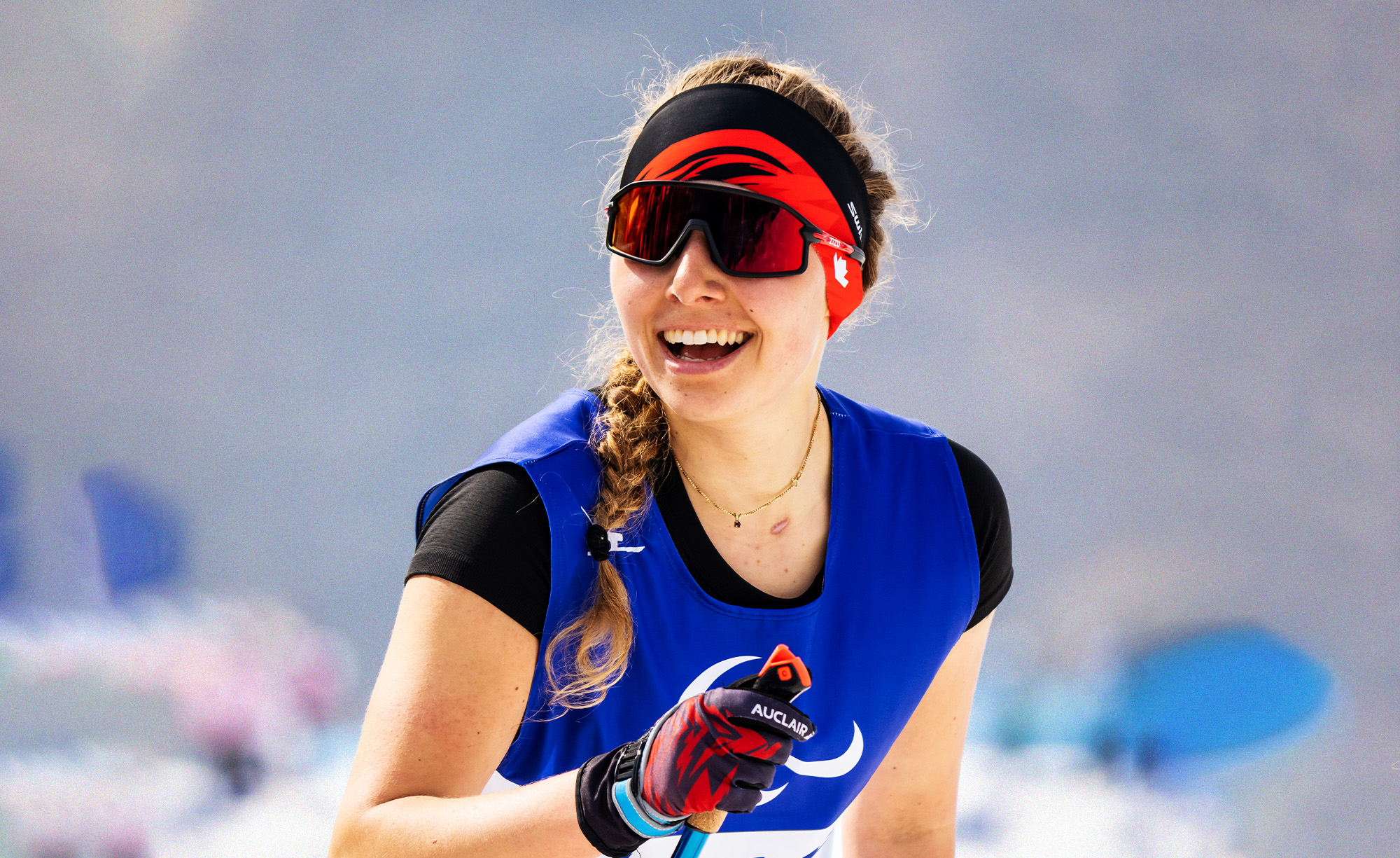 Canadian Paralympian Natalie Wilkie smiles brightly while competing in a Para Nordic skiing event. She wears a blue race bib, red headband, and sunglasses, with her ski pole in hand. The snowy background is blurred.