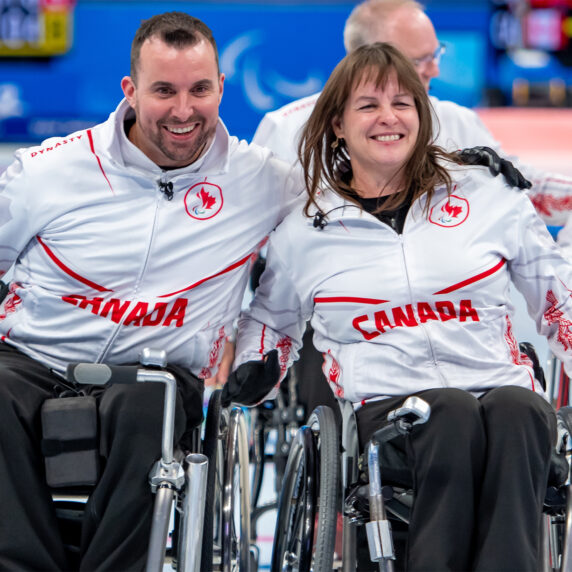 Two Canadian wheelchair curling athletes smile together on the ice, wearing white jackets with red maple leaf accents and the word "CANADA" printed across their chests. Teammates are visible in the background during a curling match.