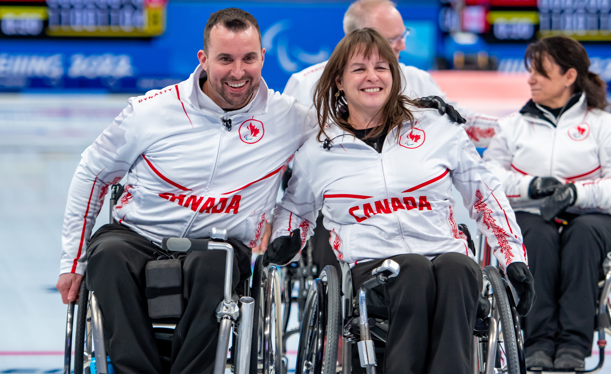 Two Canadian wheelchair curling athletes smile together on the ice, wearing white jackets with red maple leaf accents and the word "CANADA" printed across their chests. Teammates are visible in the background during a curling match.