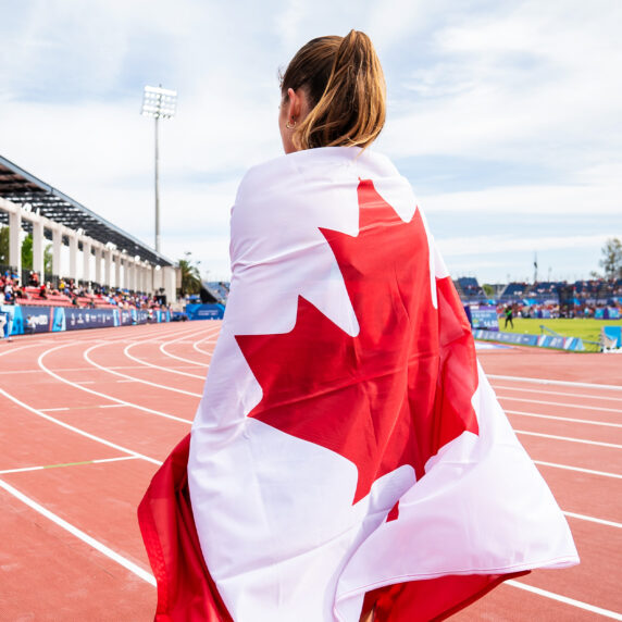 Canadian Paralympian Keegan Gaunt draped in the Canadian flag, standing on the track during the Santiago 2023 Parapan American Games. The stadium is filled with spectators under a bright sky.