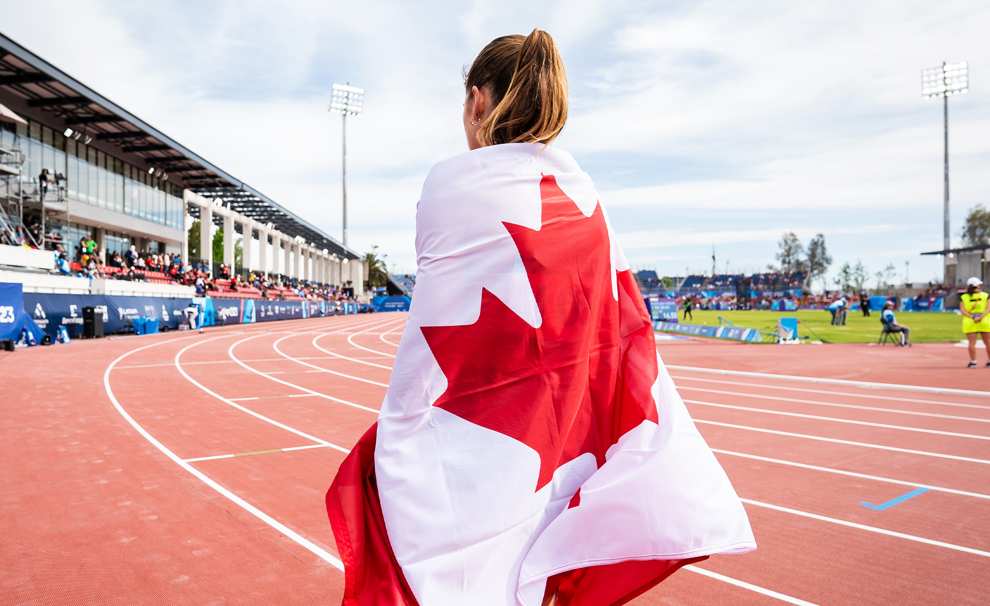 Canadian Paralympian Keegan Gaunt draped in the Canadian flag, standing on the track during the Santiago 2023 Parapan American Games. The stadium is filled with spectators under a bright sky.