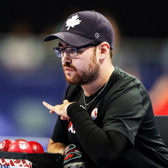 Canadian Boccia Paralympian Danik Allard focuses intently during a match. Wearing a black shirt and a black cap with a white maple leaf, he is seated next to red Boccia balls in a Canada-themed box. The background is blurred with vibrant colours.