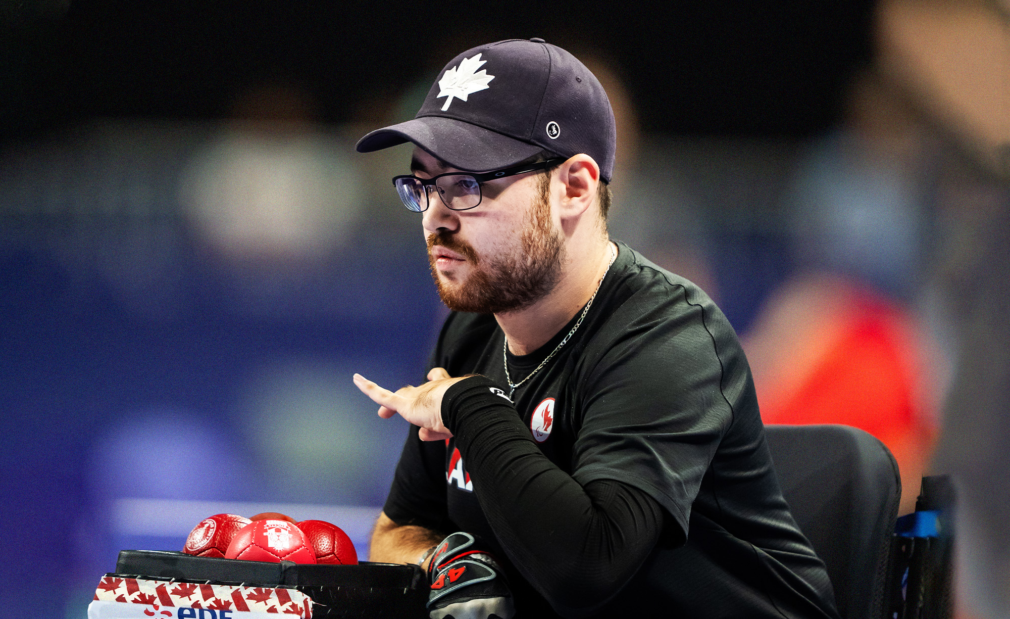 Canadian Boccia Paralympian Danik Allard focuses intently during a match. Wearing a black shirt and a black cap with a white maple leaf, he is seated next to red Boccia balls in a Canada-themed box. The background is blurred with vibrant colours.