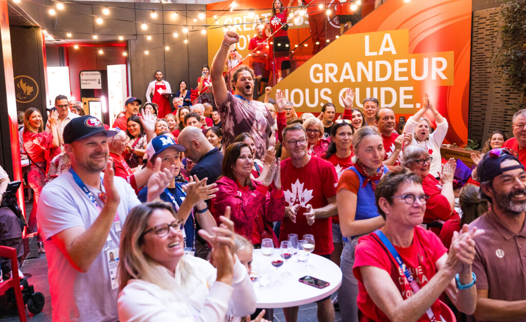 A vibrant crowd of Canadian fans dressed in red and white cheer enthusiastically at the Canadian Paralympic House event during the Paris 2024 Paralympics. The venue is adorned with Canadian branding, including a banner that reads 'La Grandeur Nous Guide.' The atmosphere is festive, with string lights overhead and smiles all around.