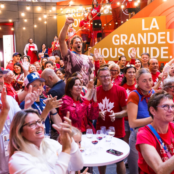 A vibrant crowd of Canadian fans dressed in red and white cheer enthusiastically at the Canadian Paralympic House event during the Paris 2024 Paralympics. The venue is adorned with Canadian branding, including a banner that reads 'La Grandeur Nous Guide.' The atmosphere is festive, with string lights overhead and smiles all around.