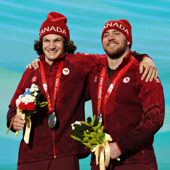 Mac Marcoux (right) stands on the podium at the Beijing 2022 Paralympic Games, wearing a red Team Canada jacket and toque, holding a bouquet of flowers. He smiles alongside his guide, Tristan Rodgers (left), as they celebrate winning the silver medal in the Men's Downhill, visually impaired event.