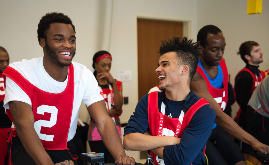 Two young men wearing red numbered pinnies laugh together while participating in an indoor cycling activity. In the background, other participants in similar attire are engaged in activities, creating a lively and energetic atmosphere.
